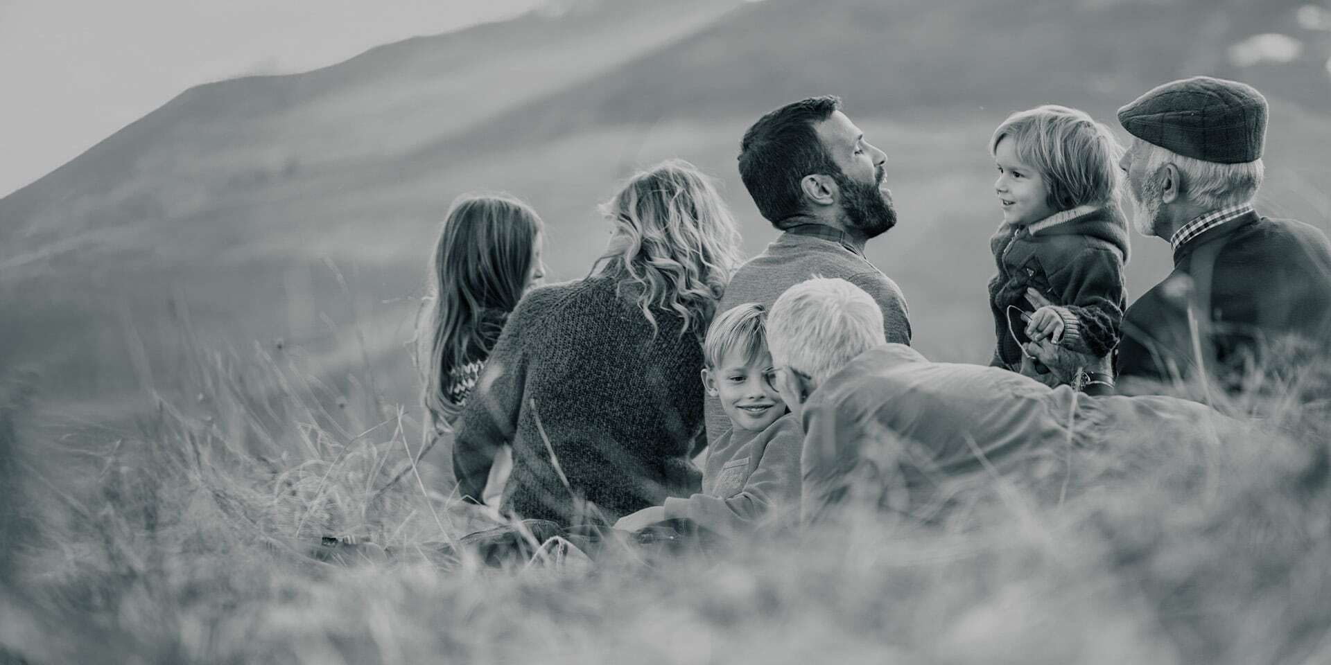 A multi-generational family sitting together on the grass, talking and laughing.
