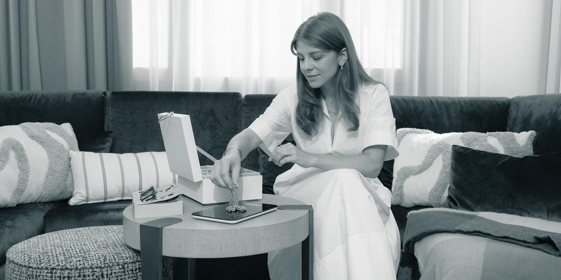 A young woman in an elegant dress sits on a modern couch, weighing gold jewellery she takes from a jewellery box.