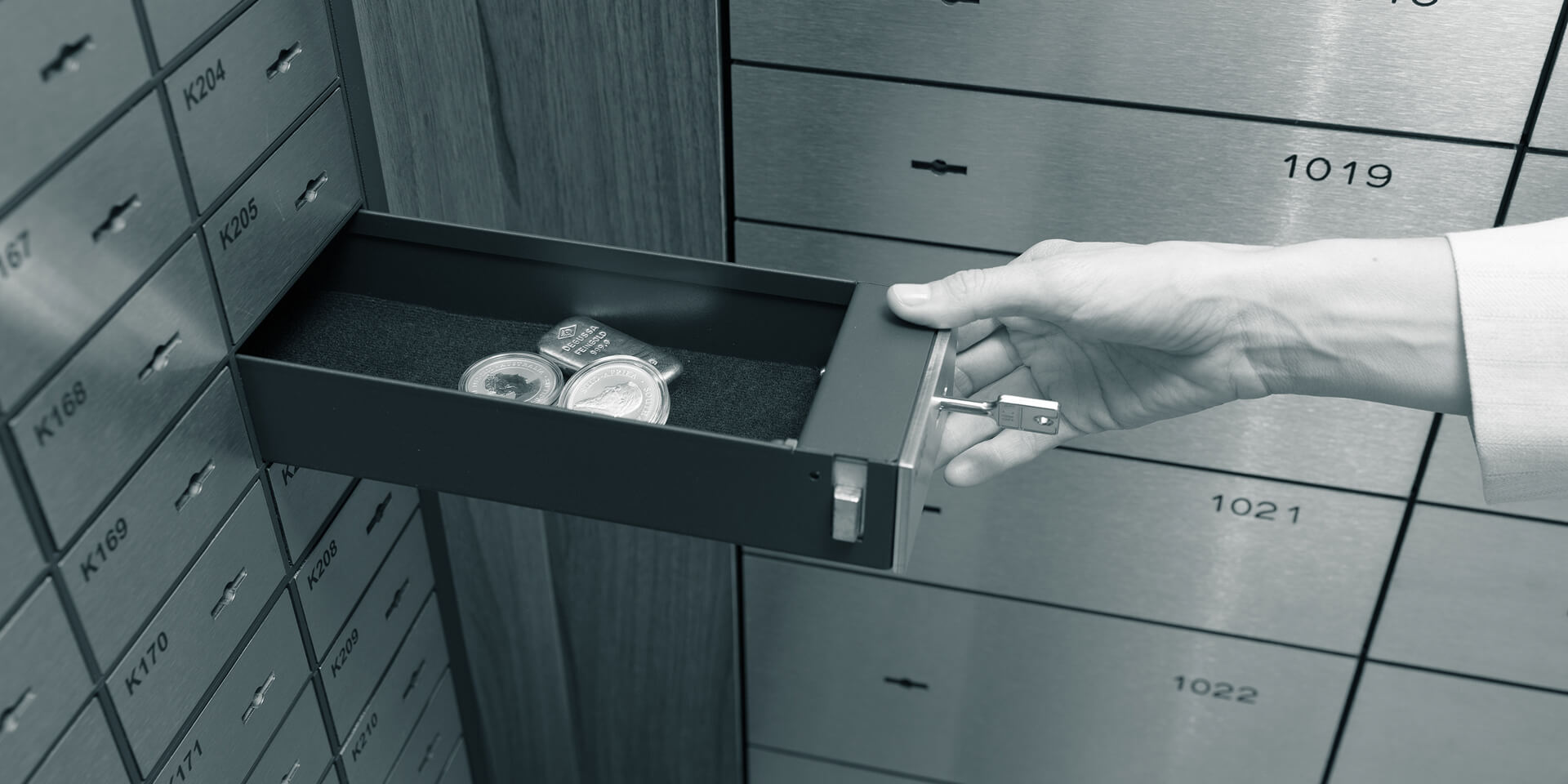 A hand opens a safe deposit box drawer in a room full of safe deposit boxes, holding coins and a gold bar.